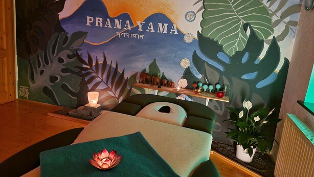 Una habitación con un mural de paisaje tropical, una camilla de masaje con decoración de flor de loto y un estante con adornos.