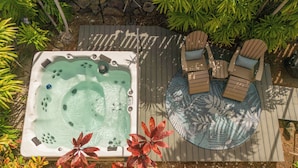 A hot tub with a wooden deck, surrounded by tropical plants and a seating area with chairs and a table. - Kona Polynesian King - Tropical Island Soul (Kailua-Kona)