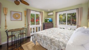 A bedroom with a bed, a desk, chairs, a ceiling fan, and large windows with curtains. - Kona Polynesian King - Tropical Island Soul (Kailua-Kona)