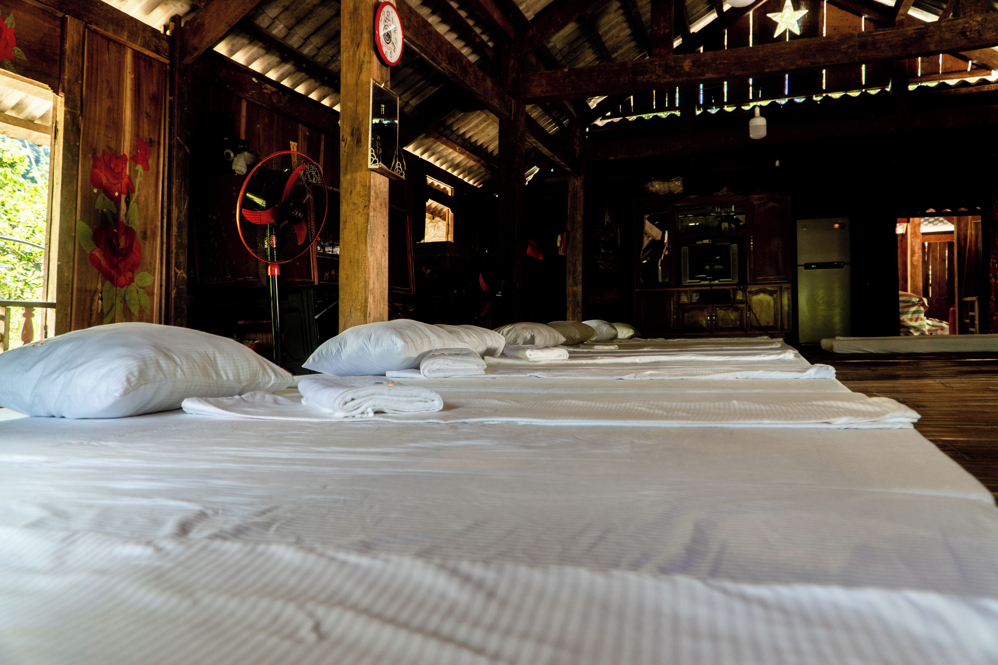 A row of neatly made beds with white linens in a rustic room with wooden beams and a tiled ceiling.