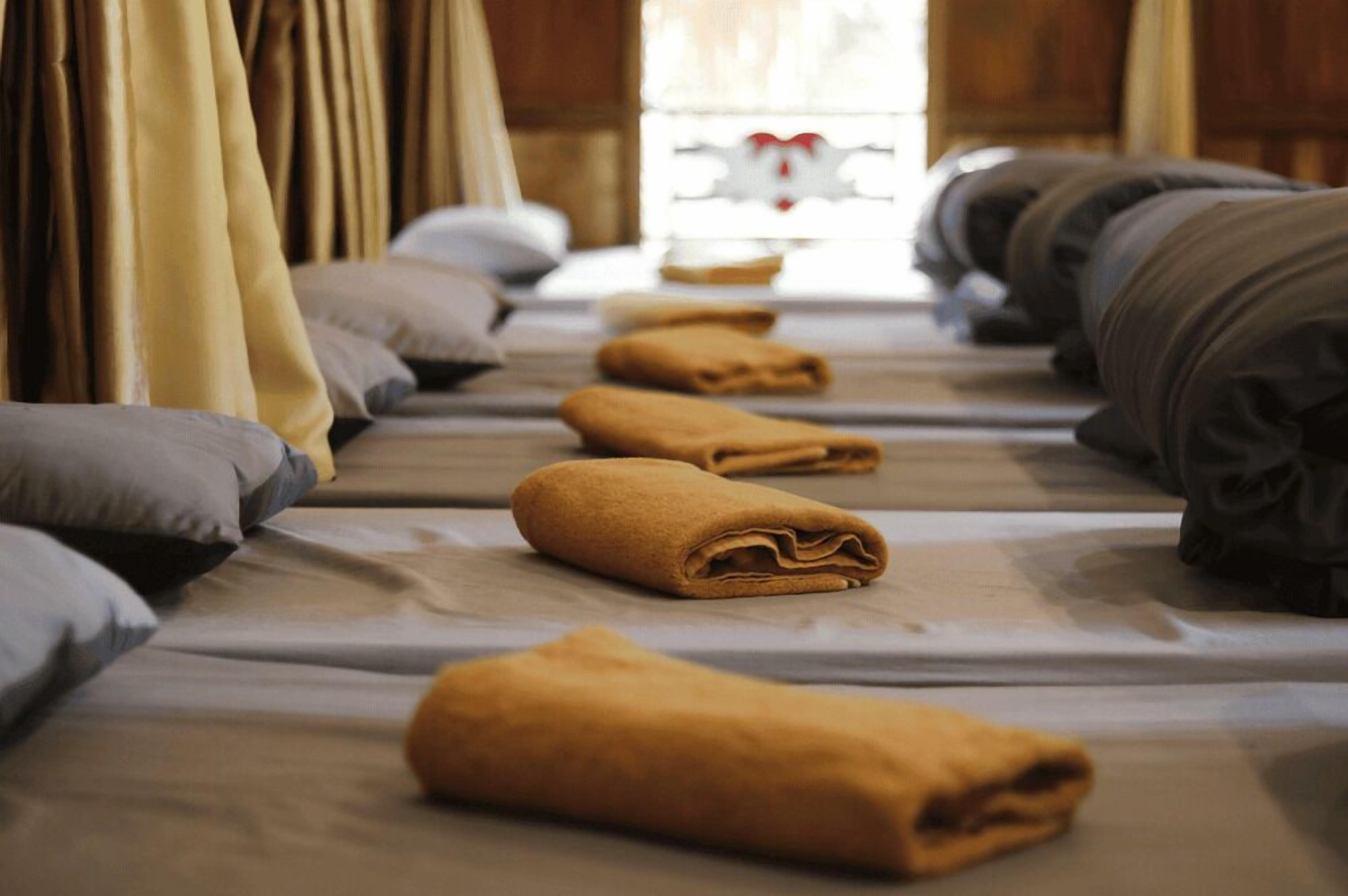 A row of neatly made beds with folded towels, a wooden floor, and a window with curtains.
