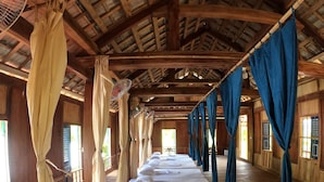 Una habitación con vigas de madera, una hilera larga de camas con ropa blanca y cortinas en tonos beige y azul.