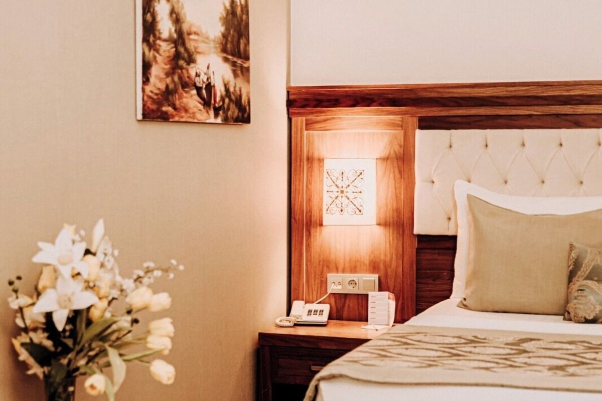 Habitación de hotel con cama, mesita de noche con lámpara, un jarrón de flores y un cuadro en la pared.
