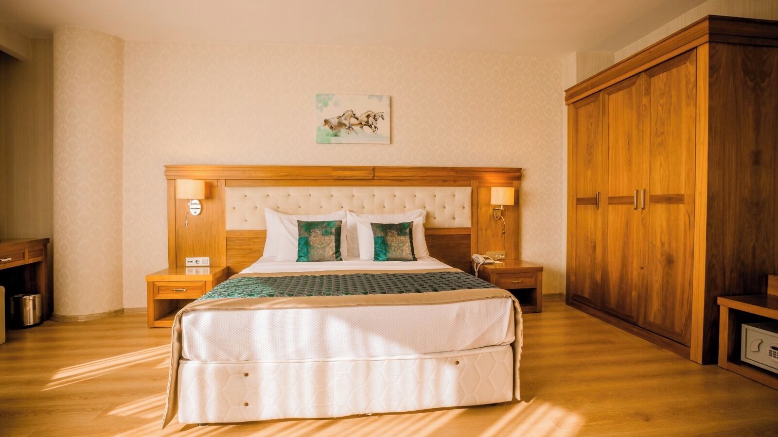 A hotel room with a large bed, wooden headboard, two bedside tables with lamps, a wooden wardrobe, and a painting on the wall.