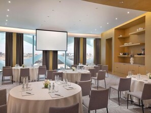 Un salón de conferencias con varias mesas, sillas, una pantalla de proyector y vistas a la ciudad.