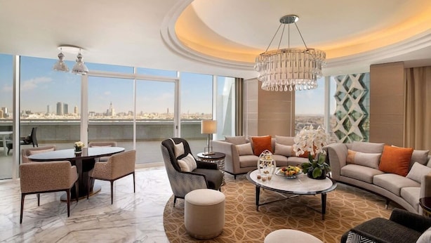A modern living room with a round coffee table, sofas, a dining area with a table and chairs, and a large window offering a city view. - Sofitel Riyadh Hotel & Convention Centre (Riyadh)