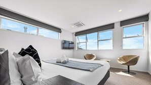 A modern hotel room with a large bed, a small desk, two chairs, and large windows offering a view of the sky. - Orakei Oasis l Quality 3 Bed House inc. Views (Auckland)