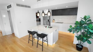 A modern kitchen with white cabinetry, a central island with bar stools, pendant lighting, and a potted plant. - Orakei Oasis l Quality 3 Bed House inc. Views (Auckland)