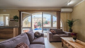 A modern living room with a sofa, a coffee table, and a view of a swimming pool and garden through large glass doors. - Ellie Boo - Farmhouse w/ Pool, Hot Tub, Games Room (Xewkija)