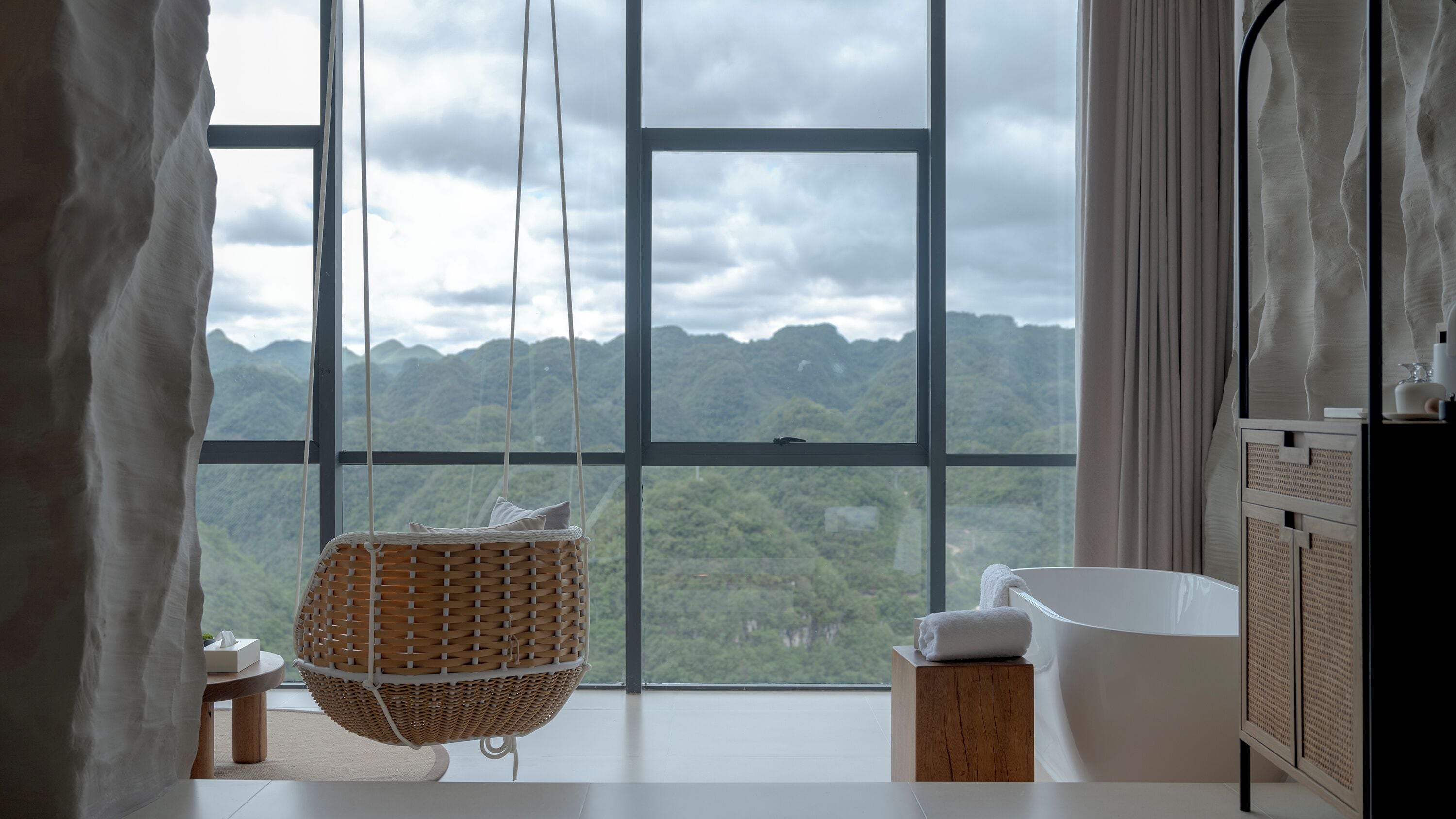 A bathroom with a large window offering a mountain view, a hanging wicker chair, and a white bathtub.