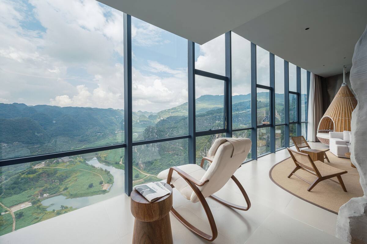 A modern interior with large windows offering a view of a mountainous landscape. There are two rocking chairs and a small wooden table.