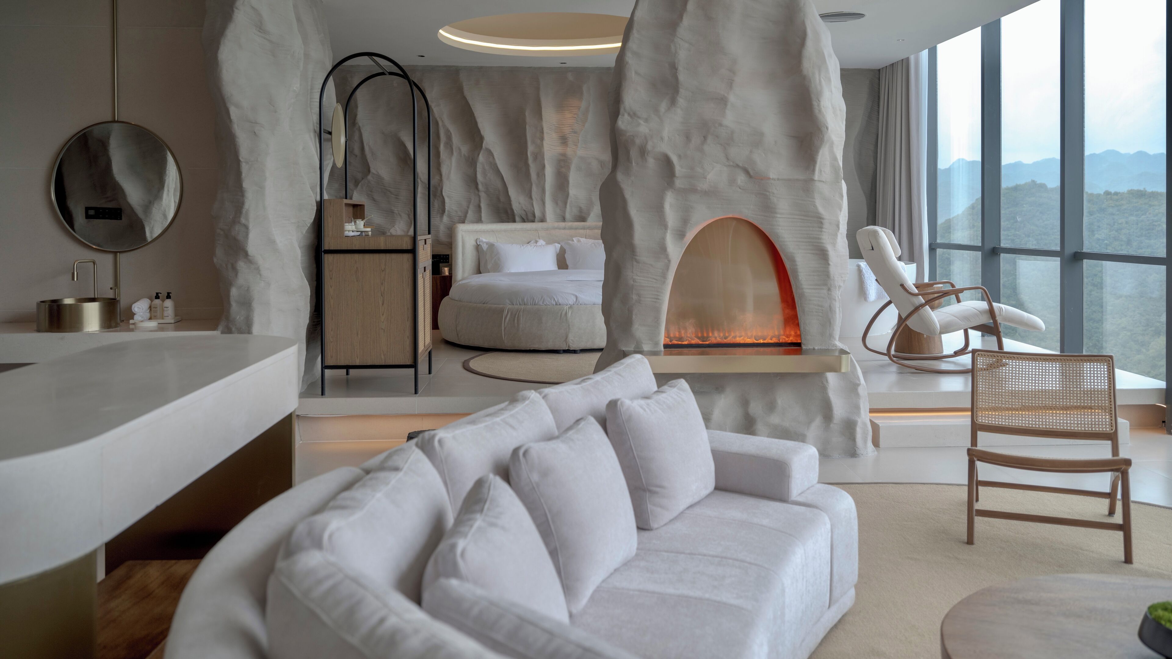 A modern hotel room with a large window, a fireplace, a sofa, and a wooden cabinet.