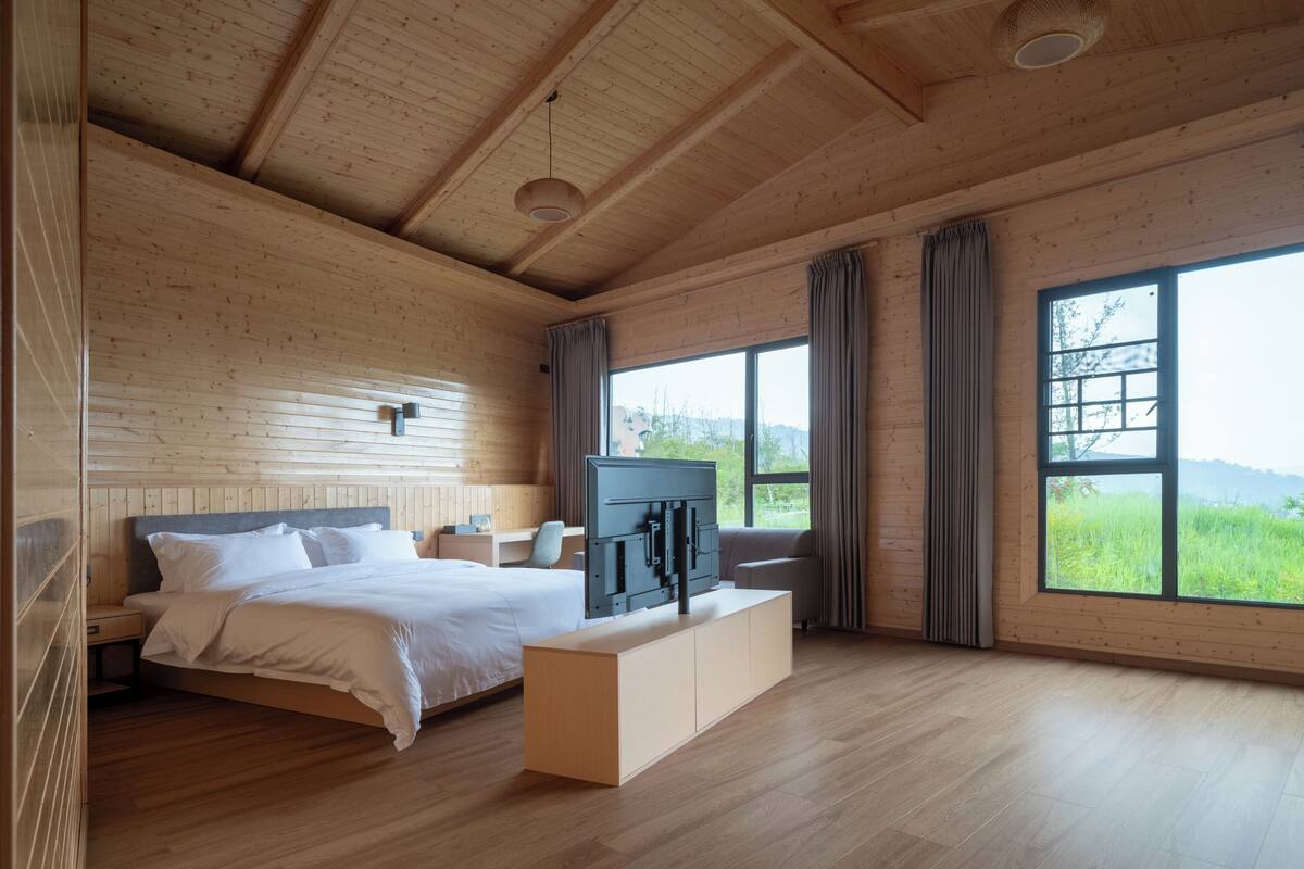 A modern bedroom with a wooden ceiling, a large bed, a TV, and a view of greenery outside.