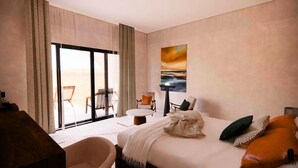 Une chambre d’hôtel avec un lit, un bureau, une chaise, une petite table et une vue sur la plage, visible à travers une grande fenêtre.