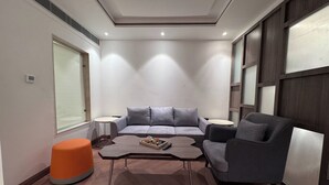 A modern living room with a grey sofa, a black armchair, and an orange ottoman. There is a wooden coffee table with a tray of items. The room has a herringbone floor pattern and large windows on one side. - Zone Connect by The Park Prayagraj (Prayagraj)