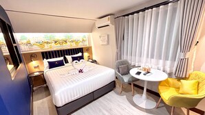 A hotel room with a bed, a small round table, a yellow chair, a gray armchair, and a view of a garden through the window.