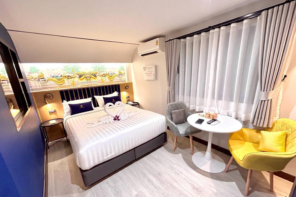Una camera d'hotel con un letto, un tavolino rotondo, una sedia gialla, una poltrona grigia e vista su un giardino dalla finestra.