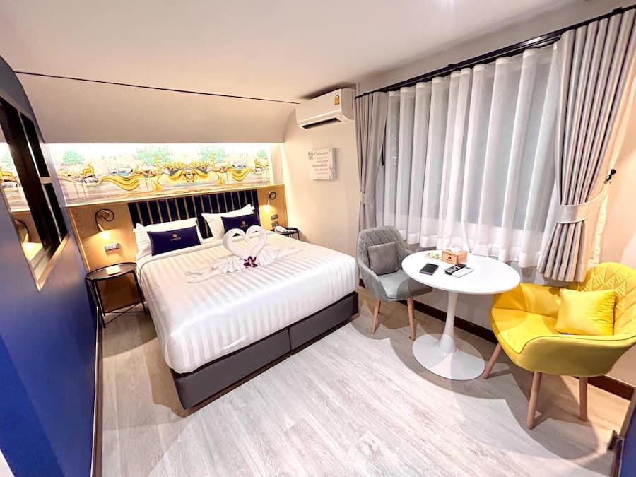 A hotel room with a bed, a small round table, a yellow chair, a gray armchair, and a view of a garden through the window.