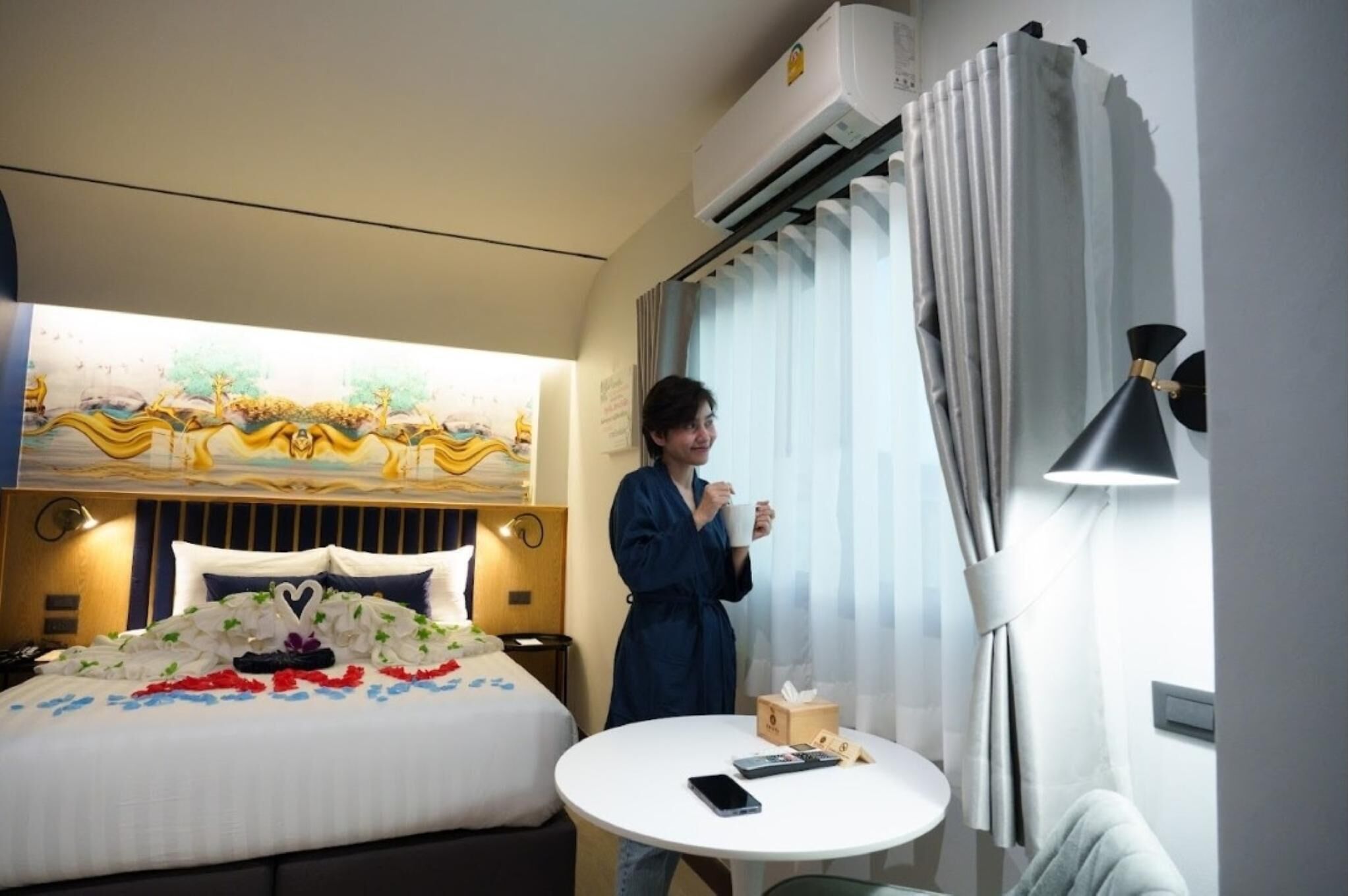 Une chambre d’hôtel avec un lit, une table ronde, un lampadaire et une fenêtre avec des rideaux.