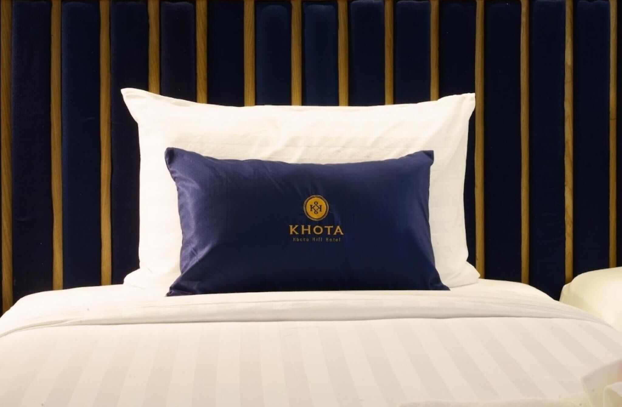 A bed with a white and navy blue pillow featuring the Khota Hotel logo.