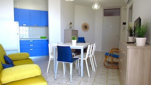 A modern kitchen with blue cabinets, a dining area with a white table and blue chairs, a yellow sofa, and a wooden cabinet with plants. - Apartment in Caorle Near Beach & Pool (Caorle)