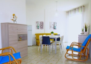 A dining area with a white table, blue chairs, a yellow sofa, and a wooden cabinet. - Apartment in Caorle Near Beach & Pool (Caorle)