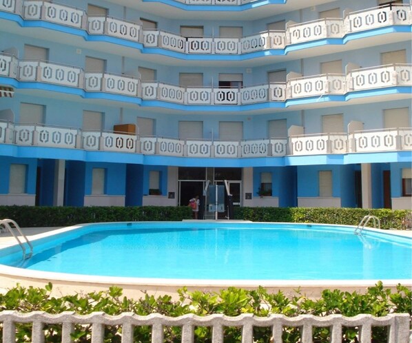 A multi-story hotel with a swimming pool, balconies with geometric patterns, and a person entering the building. - Apartment in Caorle Near Beach & Pool (Caorle)