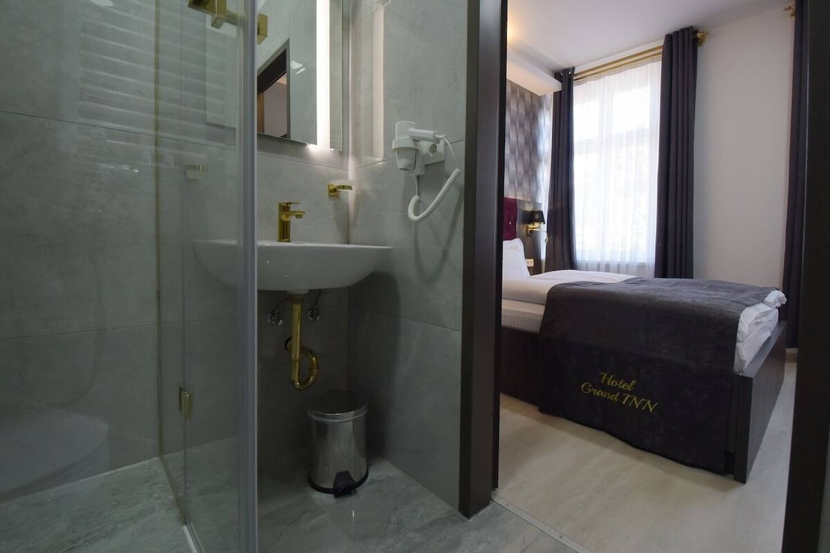 Ein Hotelzimmer mit einer gläsernen Duschkabine, einem weißen Waschbecken mit goldfarbenen Armaturen, einem Bett mit dunkler Bettdecke und einem Fenster mit Vorhängen.