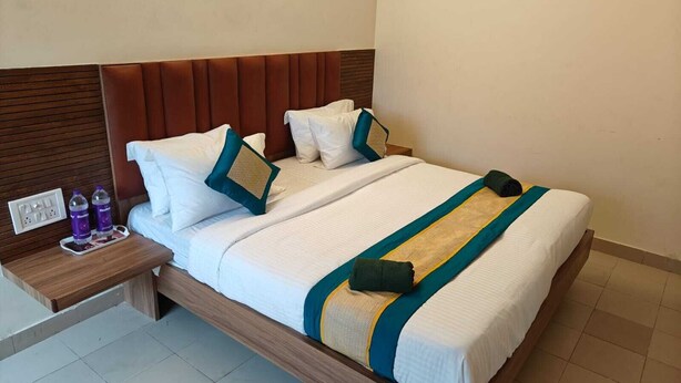 A neatly made bed with white linens and teal and yellow accents, a wooden headboard, and a bedside table with bottled water. - Hotel GT Galaxy (Chennai)