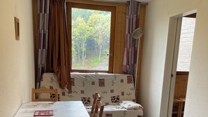 A small, cozy room with a dining table and chairs, a sofa with patterned cushions, and a window with curtains.