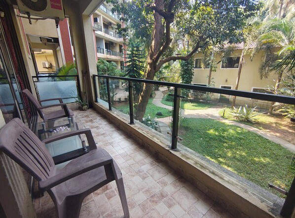 A balcony with two wooden chairs and a glass table, overlooking a garden with a pathway and a building in the background. - Tranquil 1BHK Garden Estate, Pool | Palolem Beach (Canacona)
