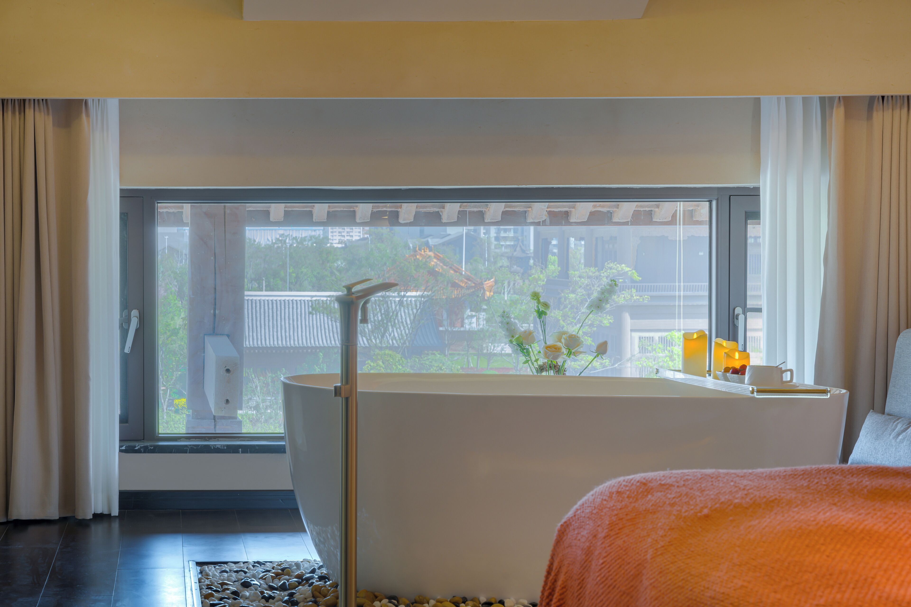 Une chambre d’hôtel moderne dotée d’une grande fenêtre, d’une baignoire blanche et offrant une vue sur un immeuble et la verdure environnante.
