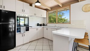 A modern kitchen with white cabinets, a black refrigerator, a built-in oven, a window with a view of trees, and a wooden ceiling.