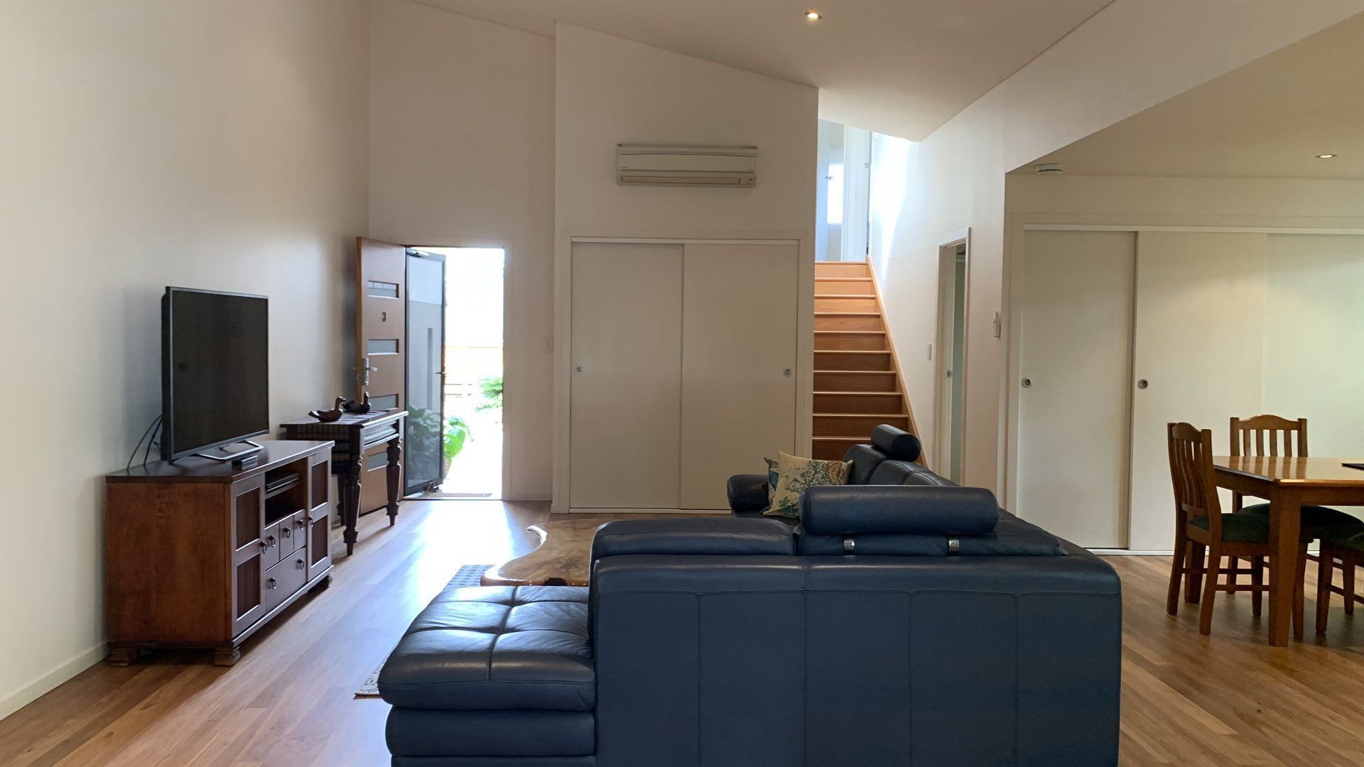 A modern living room with a sectional sofa, a television on a wooden cabinet, a dining table with chairs, and a staircase leading to an upper level.