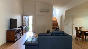 Ein modernes Wohnzimmer mit einer Eckcouch, einem Fernseher auf einer Holzkommode, einem Esstisch mit Stühlen und einer Treppe, die zu einer oberen Ebene führt.