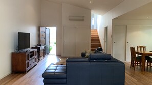 A modern living room with a sectional sofa, a television on a wooden cabinet, a dining table with chairs, and a staircase leading to an upper level. - Hook Wine & Sinker by Lifestyle. Brunswick Heads (BRUNSWICK HEADS)