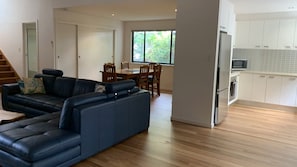 A modern living room with a sectional sofa, a dining area with wooden chairs and a table, and a kitchen with white cabinets and appliances. - Hook Wine & Sinker by Lifestyle. Brunswick Heads (BRUNSWICK HEADS)