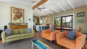 A spacious living area with a sofa, armchairs, a coffee table, and a dining table with chairs. There is a kitchen in the background with a bar counter, and a large window providing a view of the outdoors.