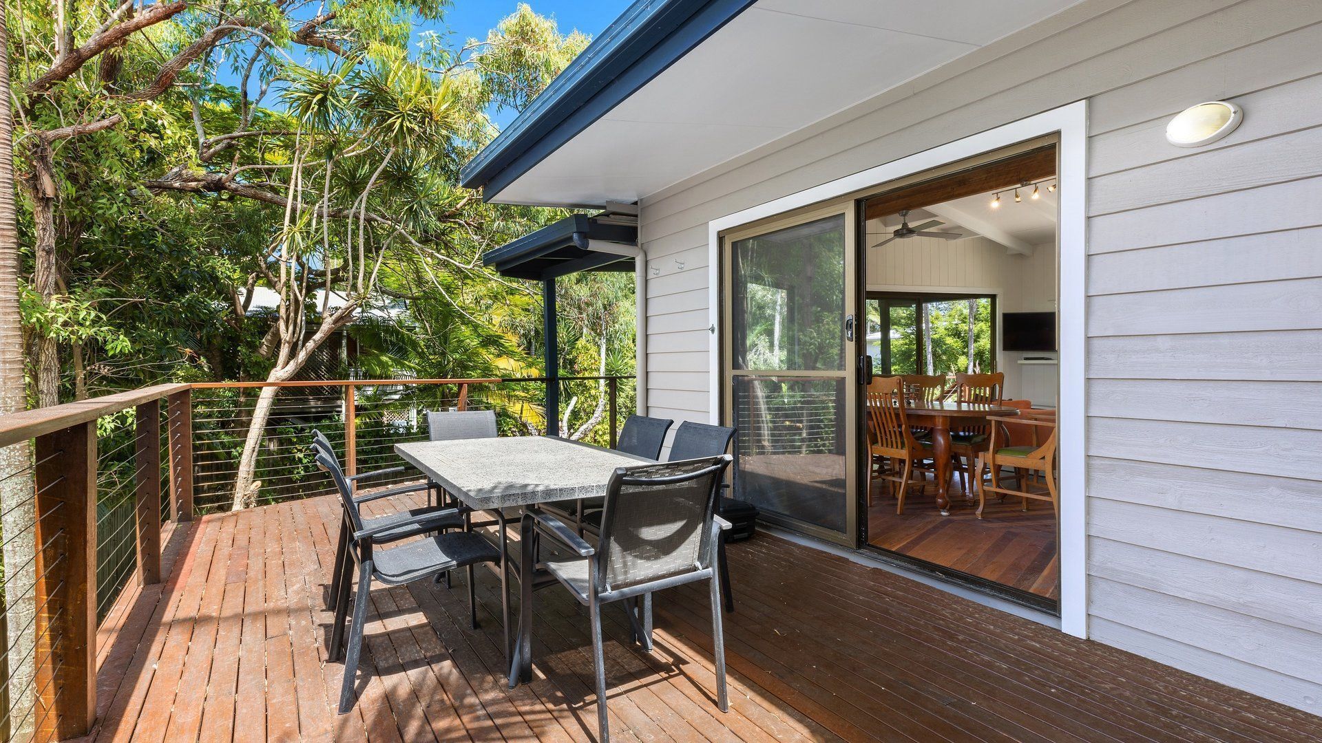 A wooden deck with a table and chairs, a glass door leading to an interior dining area, and a view of trees.