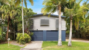 Uma casa moderna de dois andares com garagem, rodeada por vegetação exuberante e palmeiras.