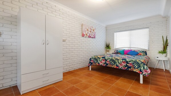 A bedroom with a white brick wall, a bed with a floral bedspread, a white wardrobe, and a window with blinds.
