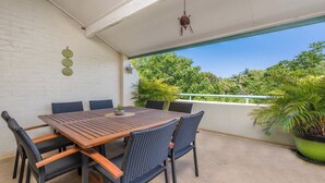 Eine überdachte Terrasse mit einem Holztisch und Korbstühlen, umgeben von Blumentöpfen und Blick auf Bäume.