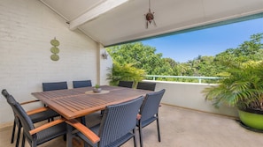 A covered patio with a wooden table and wicker chairs, surrounded by potted plants and a view of trees.