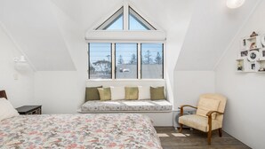 Ein Schlafzimmer mit schrägem Dach, einem großen Fenster mit Blick auf Bäume und ein Gebäude, einem Bett mit blumigem Bettbezug, einem Sessel und einem an der Wand befestigten Regal mit Bildern.