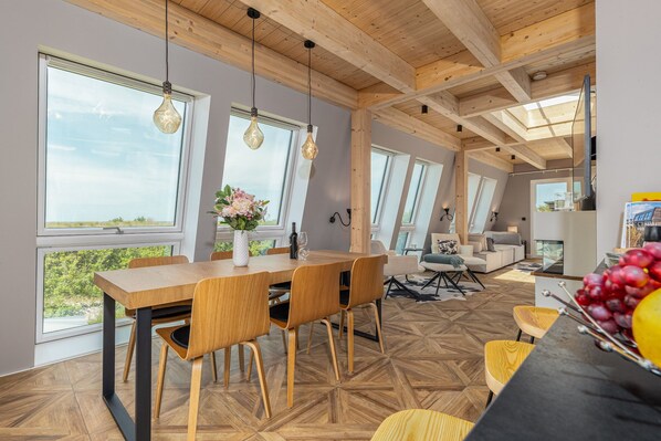 A modern dining area with a wooden table and chairs, large windows, and a view of greenery outside.