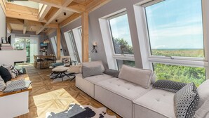 A spacious living area with a large sofa, wooden beams, and large windows offering a view of greenery.