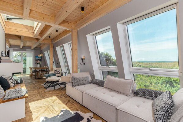A spacious living area with a large sofa, wooden beams, and large windows offering a view of greenery.