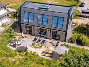 Ein modernes, zweistöckiges Gebäude mit flachem Dach, großen Fenstern und einer Terrasse mit Gartenmöbeln und Sonnenschirmen.