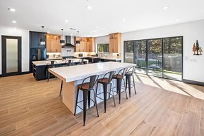 A modern kitchen with a large island, bar stools, and a view through glass doors. - The Getaway Club: Exclusive & Luxurious 7 Bed 7 Bath Retreat! Hot Tub! Pool Table! Shuffleboard! Air Hockey! Ping Pong! High Tech Theater w/ Theater Seating + Lounge Pit! Master Suite w/ Projection Screen! (Big Bear Lake)