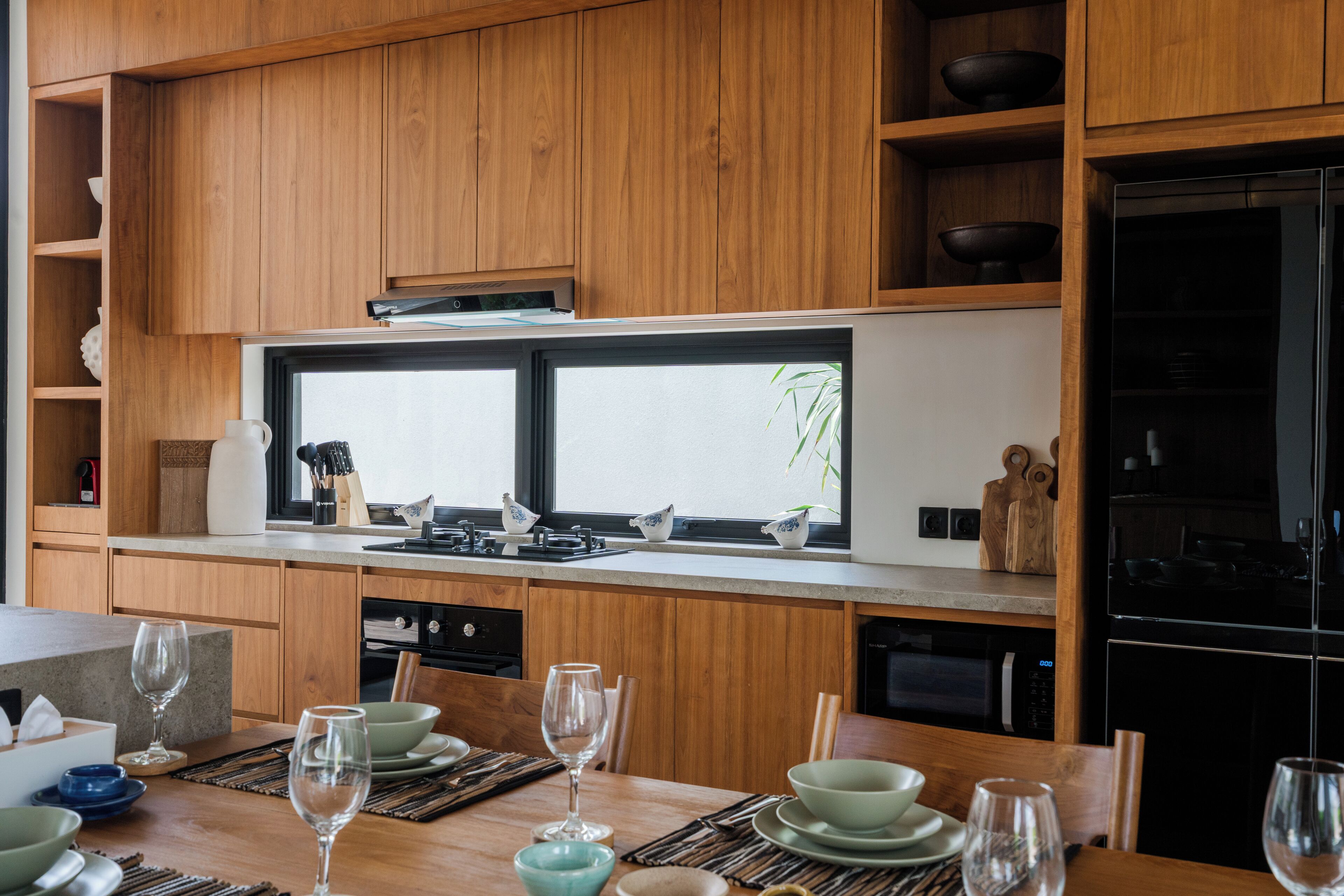 A modern kitchen with wooden cabinets, a large window, and a dining area set for six with a table, chairs, and glassware.